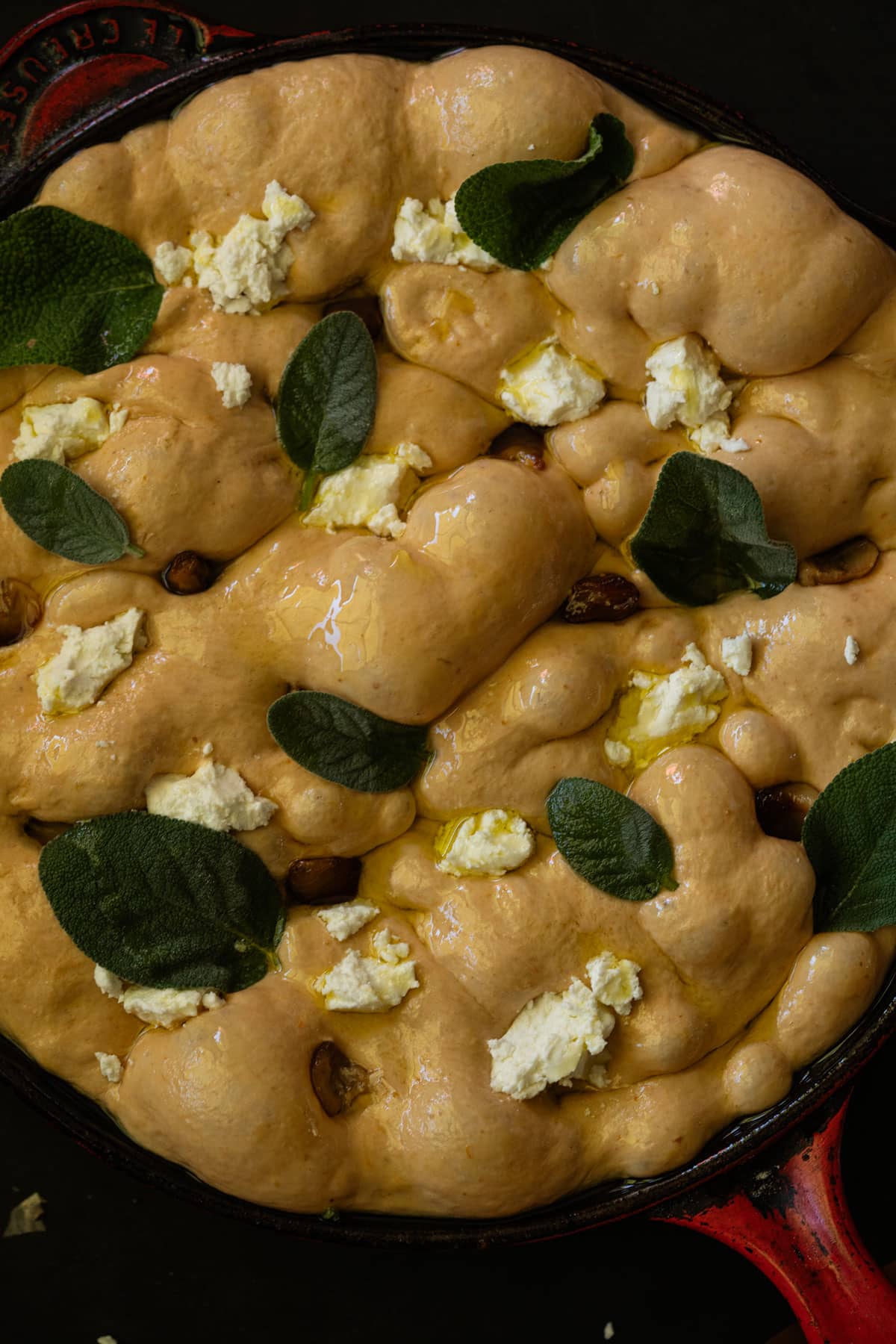 Unbaked sourdough pumpkin focaccia dough with caramelized garlic, goat cheese and fresh sage in a red skillet